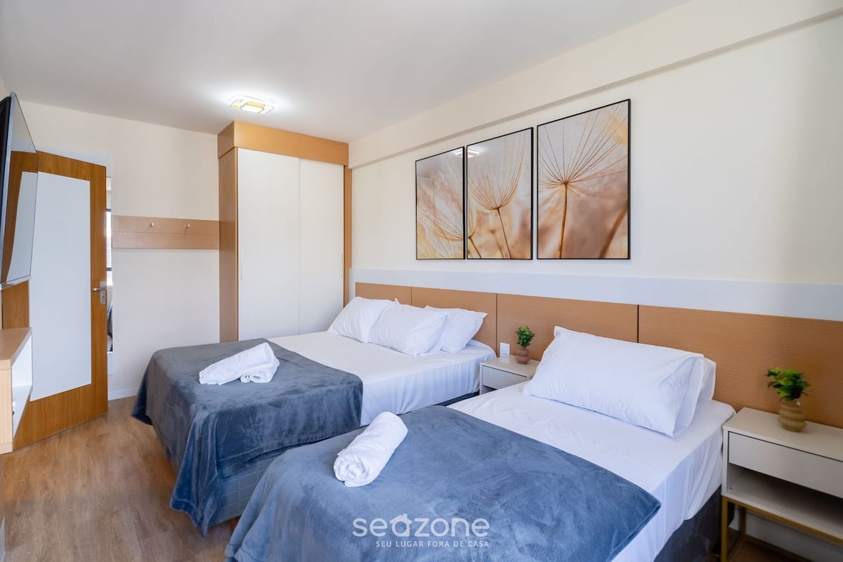 A hotel room with two beds, a wooden wardrobe, a bedside table with a plant, and a wall with three framed artworks.