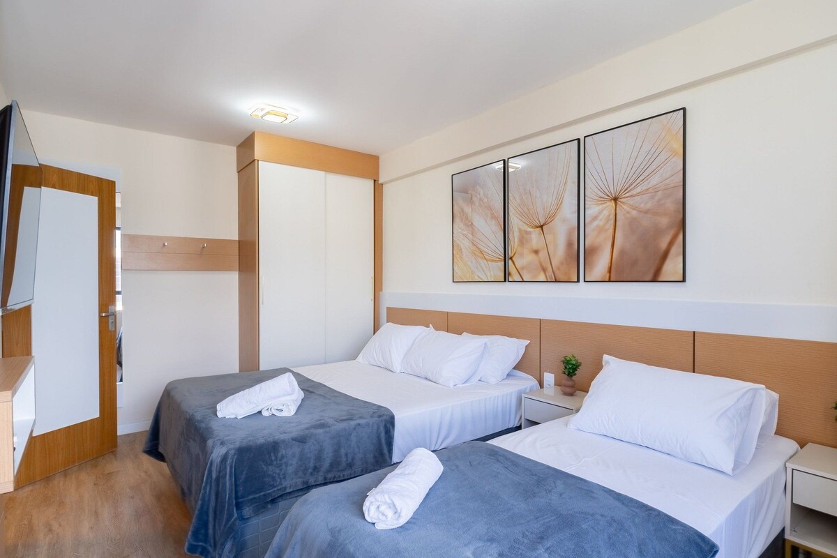 A hotel room with two beds, a wooden wardrobe, a bedside table with a plant, and a wall with three framed artworks.