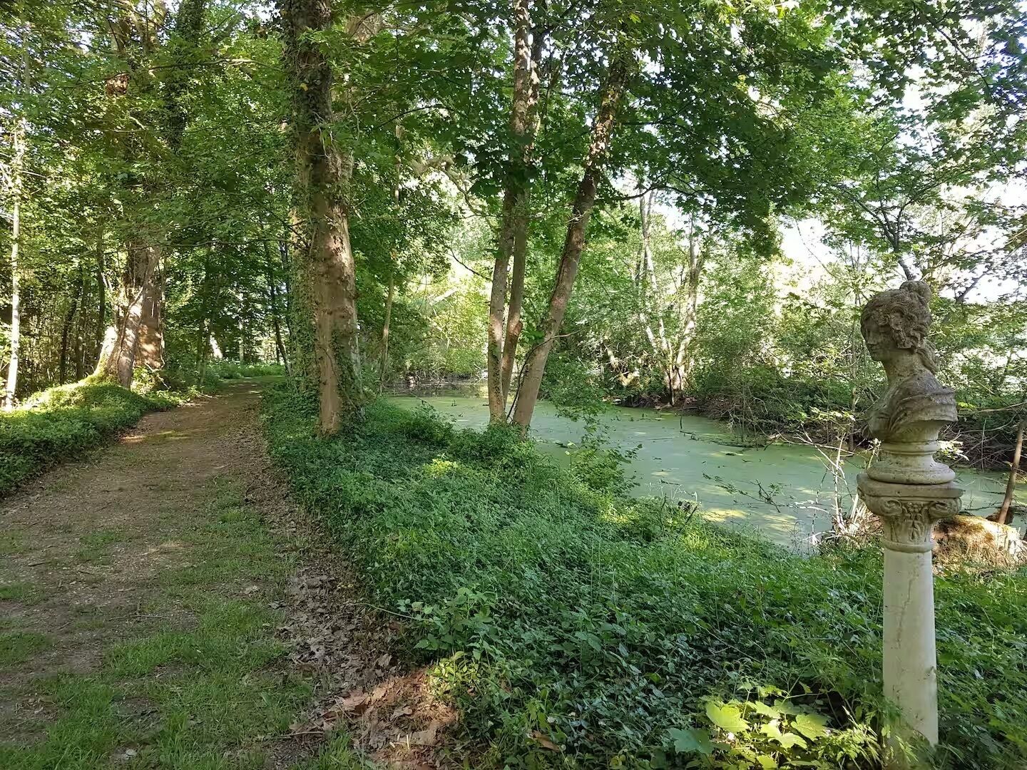 Zona boscosa con un sendero que conduce a una masa de agua, rodeada de abundante vegetación y una escultura clásica.
