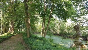 Zona boscosa con un sendero que conduce a una masa de agua, rodeada de abundante vegetación y una escultura clásica.