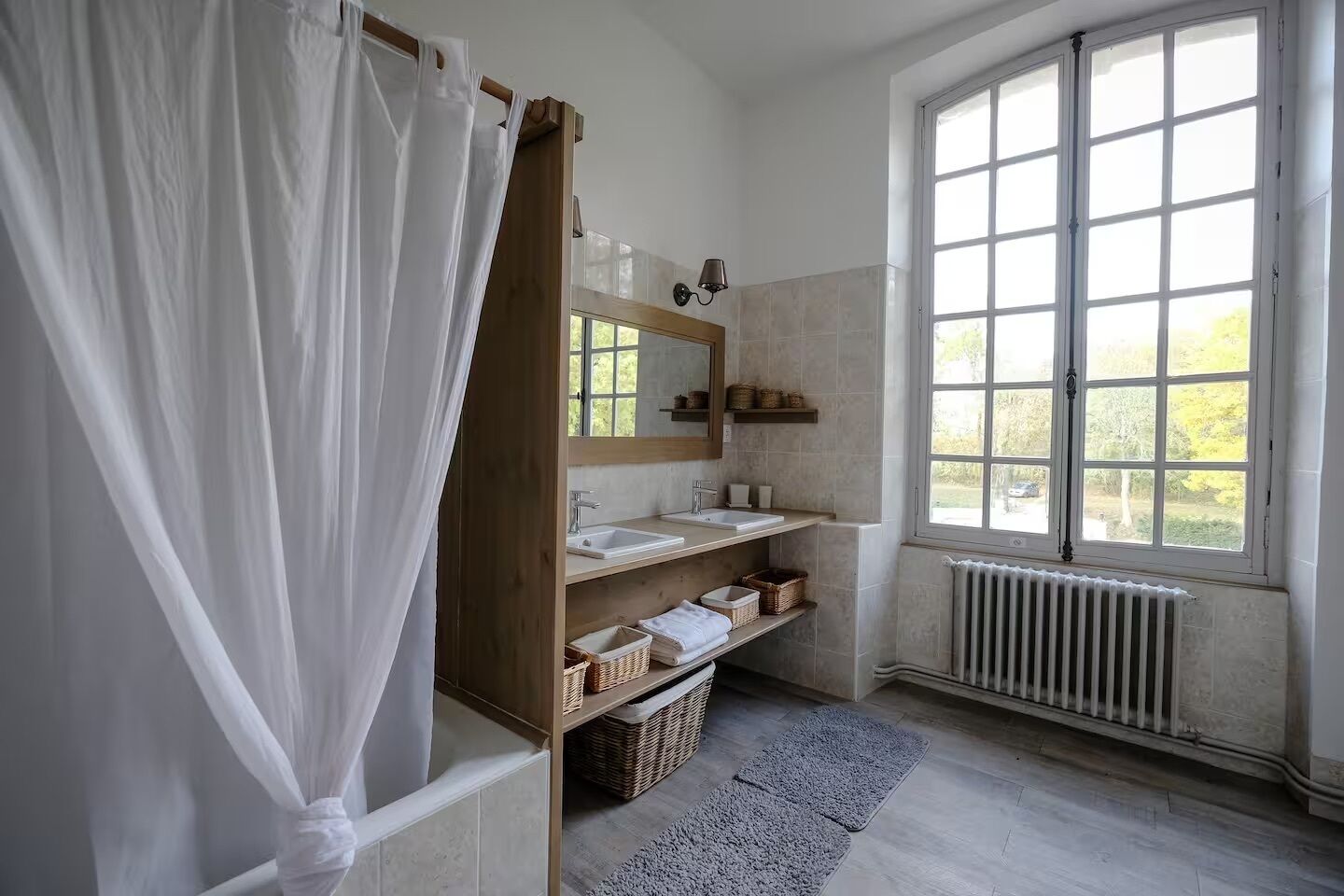 Een badkamer met een groot raam, een houten wastafelmeubel, een douche met gordijn en rieten manden.