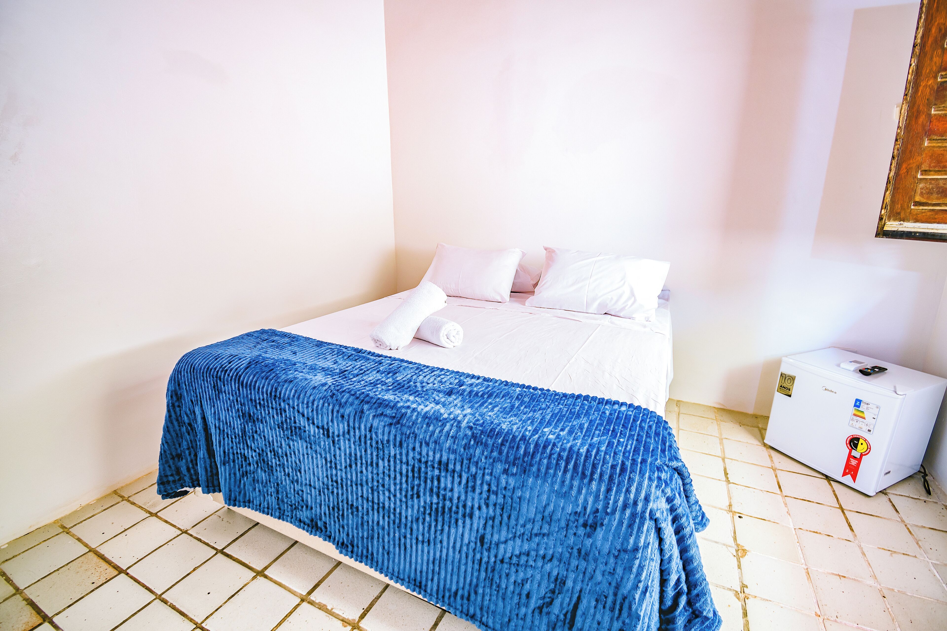 Una habitación pequeña y limpia con una cama, un edredón azul, una almohada blanca y un pequeño electrodoméstico blanco en el suelo.