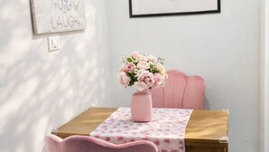Uma sala de jantar com uma mesa de madeira, cadeiras cor-de-rosa e um vaso de flores. Há duas obras de arte emolduradas na parede.