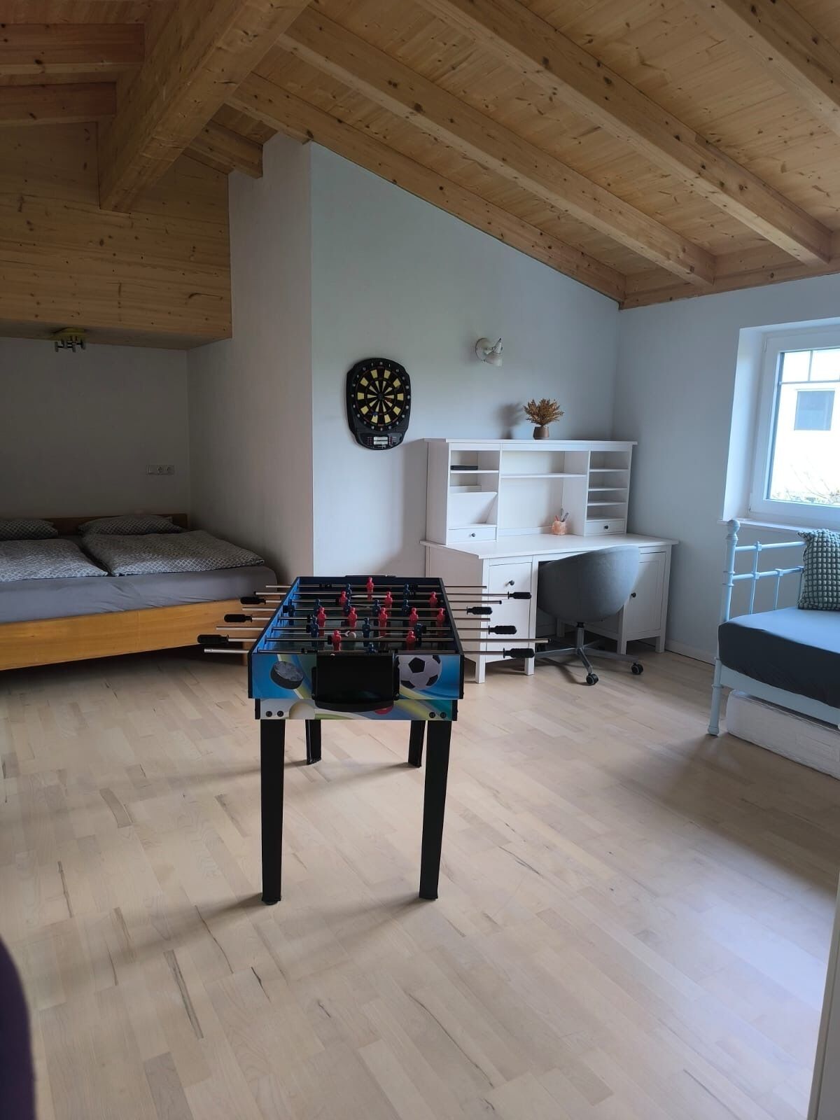 A room with a foosball table, a dartboard, a bed, a desk, and a window.