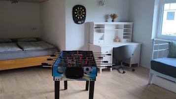 A room with a foosball table, a dartboard, a bed, a desk, and a window.