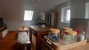 A cozy living room with a wooden floor, a dining table with chairs, a small side table, a potted plant, and a window with natural light. - 3-bedroom apartment for 6-8 persons near Munich, Augsburg (Merching)