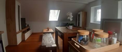 Una sala de estar acogedora con suelo de madera, una mesa de comedor con sillas, una mesita auxiliar, una planta en maceta y una ventana que deja entrar luz natural.