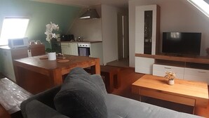 A cozy living room with a sofa, a wooden coffee table, and a TV on a wooden stand. A kitchen with a microwave, stove, and cabinets is visible in the background. - 3-bedroom apartment for 6-8 persons near Munich, Augsburg (Merching)
