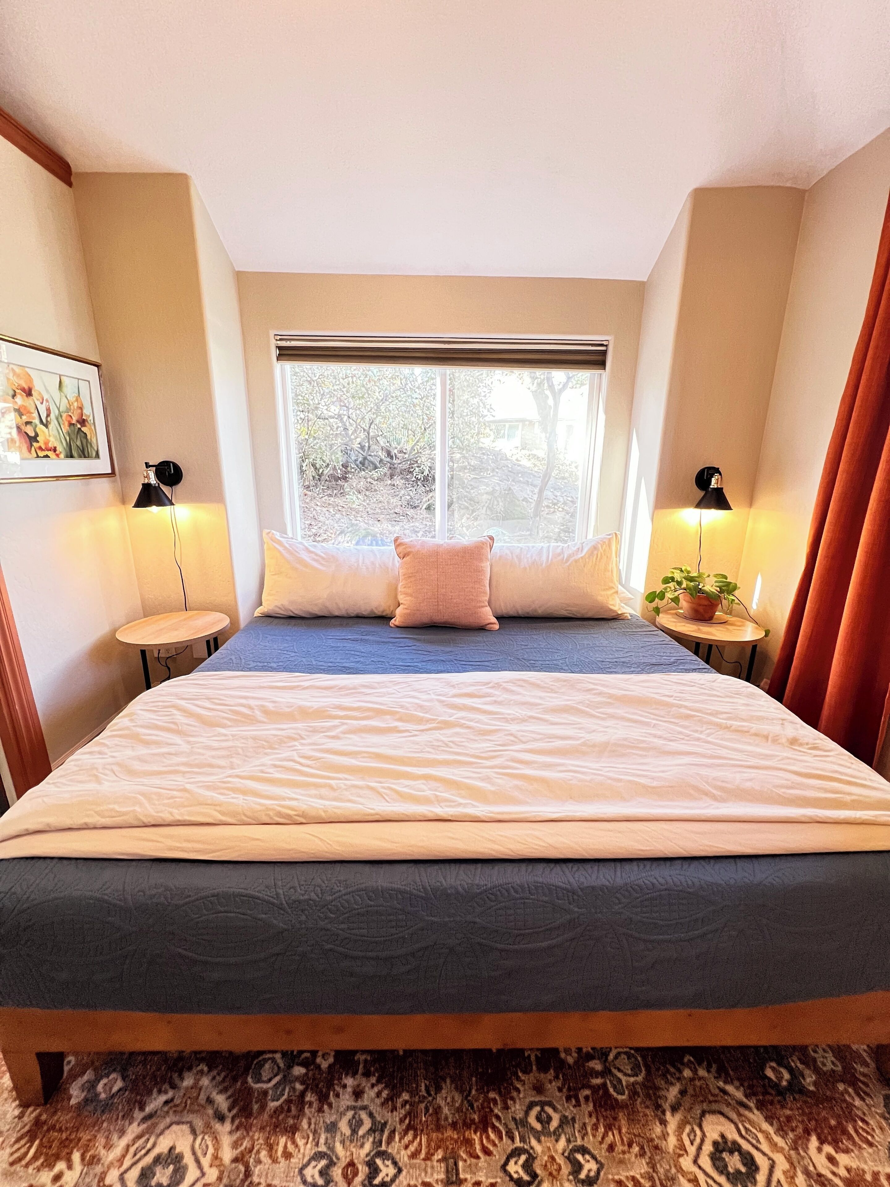 Une chambre avec un grand lit, deux tables de chevet avec des lampes, une fenêtre donnant sur des arbres et un tapis à motifs.
