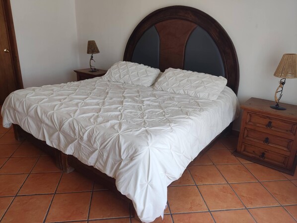 Un lit avec une couette blanche matelassée, une tête de lit en bois, une table de chevet avec une lampe et une commode en bois avec des tiroirs.