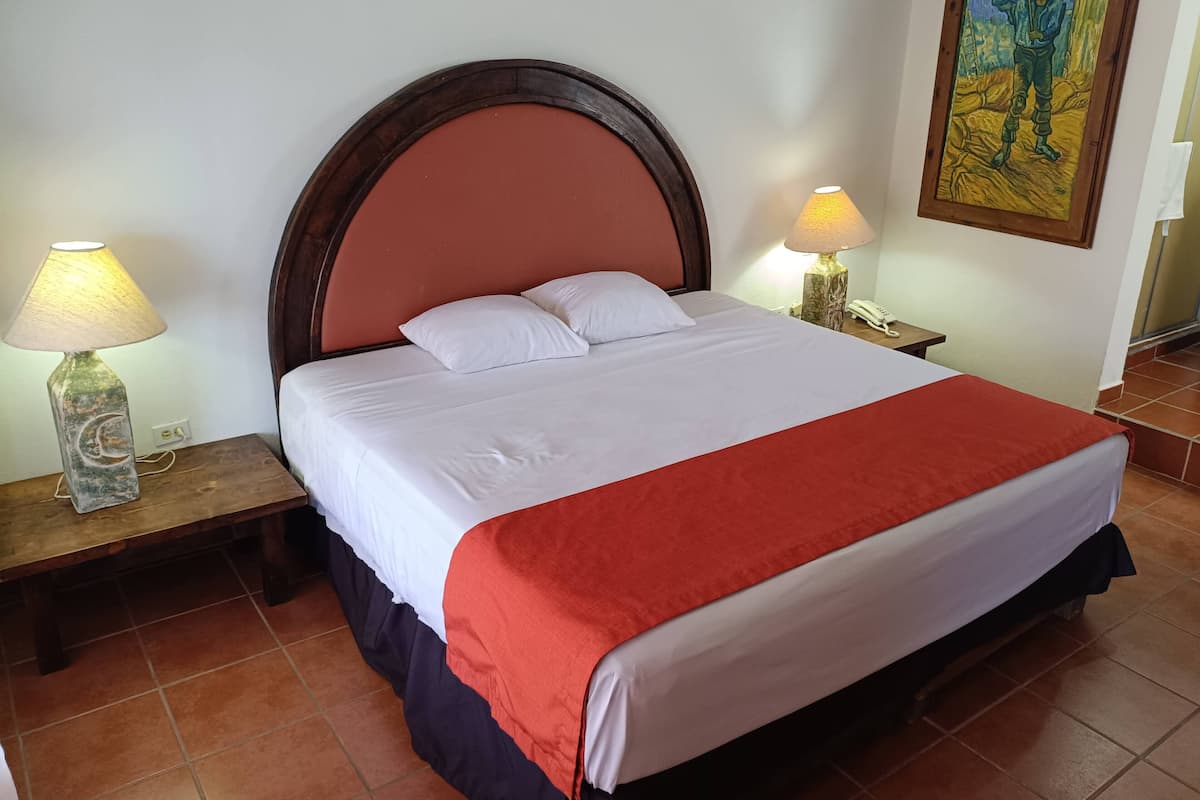 Una cama bien hecha con ropa de cama blanca y roja, dos lámparas de noche y un cuadro en la pared.