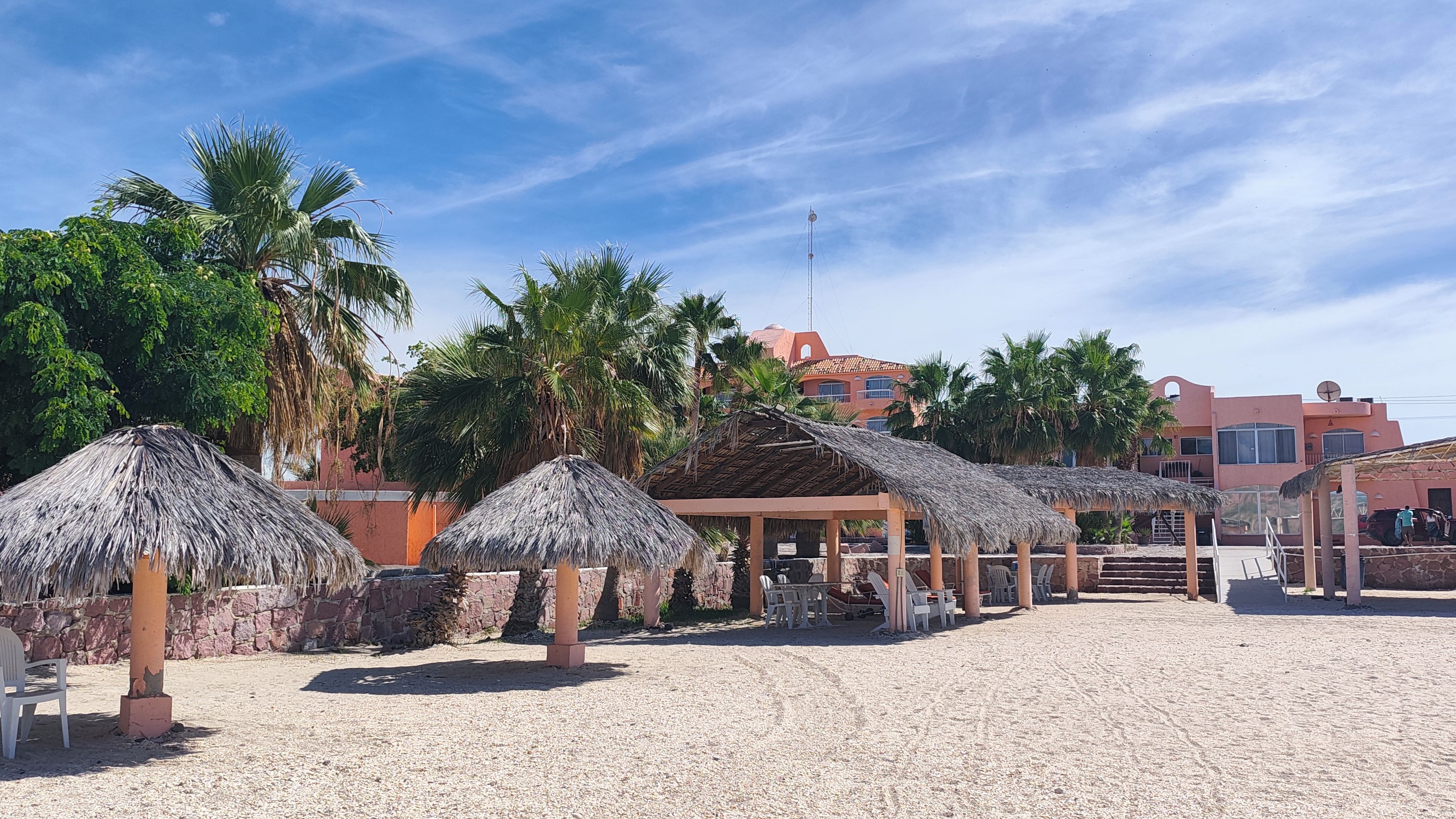 Zona de playa con sombrillas de paja, un área de estar cubierta y un edificio con fachada roja al fondo.