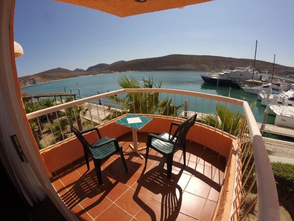 Un balcon avec une table et des chaises, donnant sur une marina où sont amarrés des bateaux, et sur un paysage vallonné.