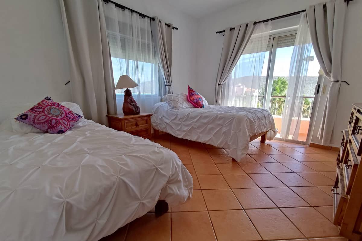 Habitación de hotel con dos camas, una mesita de noche, una lámpara y vistas al exterior.