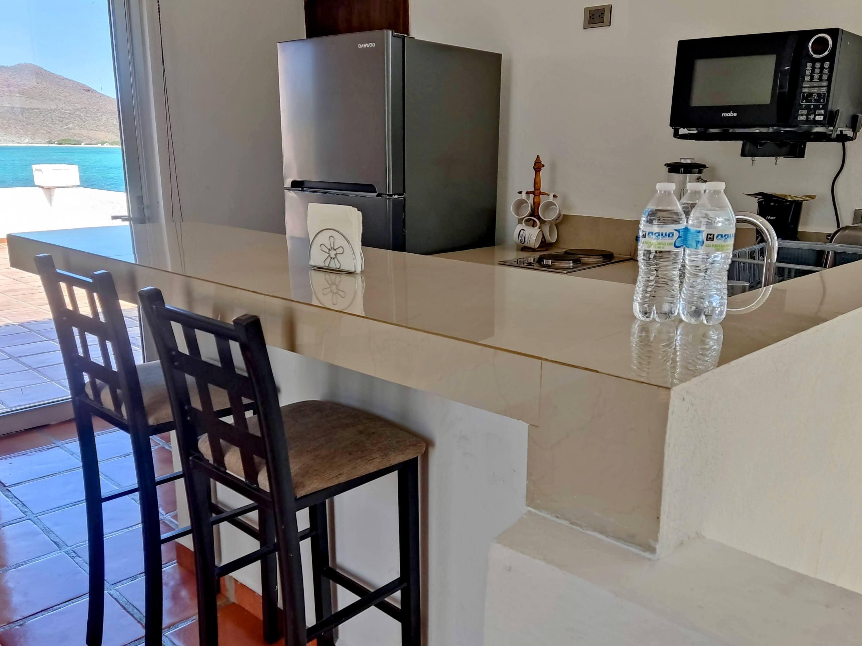Una cocina con refrigerador, microondas y una barra con sillas y botellas de agua.