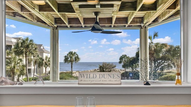 A room with a view of the ocean, a ceiling fan, a window with a 'Wild Dunes' sign, and two empty glasses on a tray. - Port O' Call D203 - Direct Ocean View! Beach! (Isle of Palms)