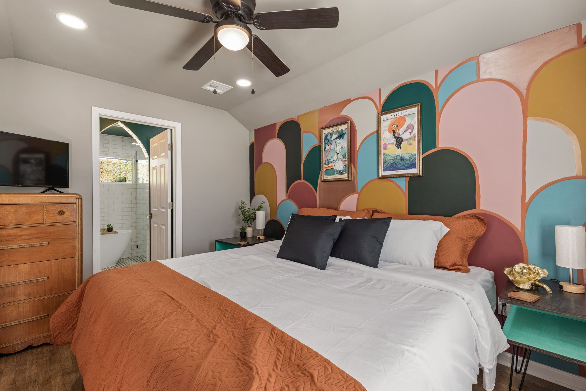 A bedroom with a bed, a dresser, a nightstand, a ceiling fan, and a wall with colorful abstract art.