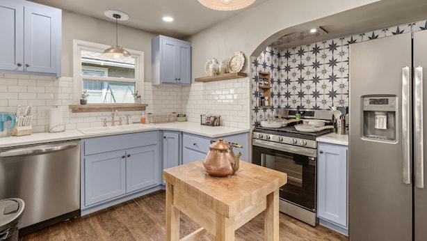 Una cocina moderna con armarios azul claro, una mesa de madera, un hervidor de cobre, un lavavajillas de acero inoxidable, un horno empotrado y un refrigerador.