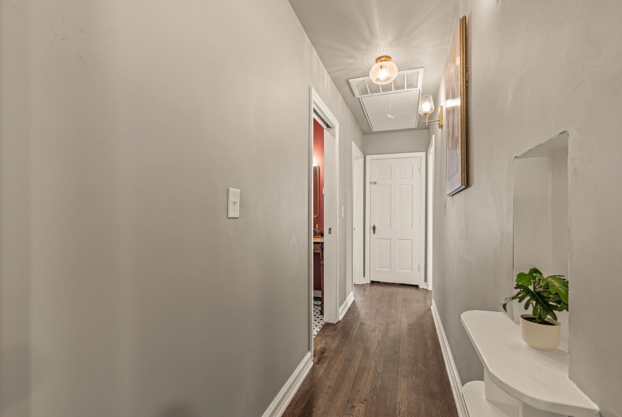 A hallway with a wooden floor, white walls, a ceiling light fixture, and a door at the end.
