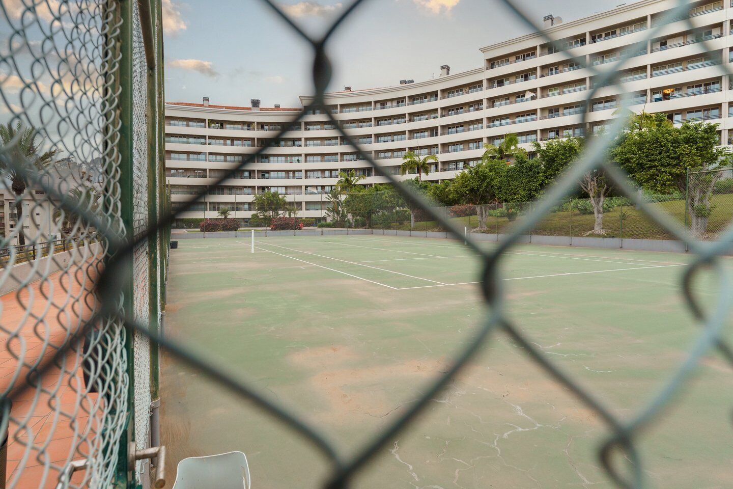 Una cancha de tenis con una cerca de malla metálica, rodeada por un edificio de varios pisos con balcones.