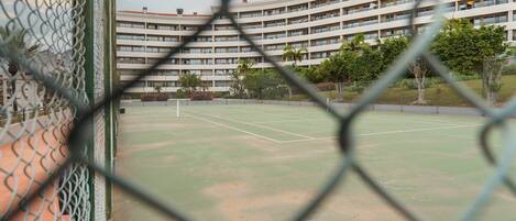 Una cancha de tenis con una cerca de malla metálica, rodeada por un edificio de varios pisos con balcones.