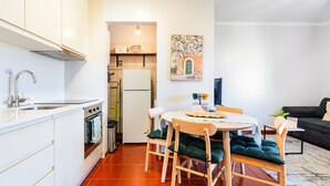 Eine kompakte Küche mit weißen Schränken, einer Edelstahl-Spüle und einem weißen Kühlschrank. Es gibt einen Essbereich mit einem runden Tisch und vier Stühlen sowie ein schwarzes Sofa im Wohnzimmer.