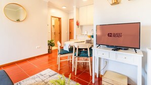 Ein modernes Wohnzimmer mit einem Flachbildfernseher, der „GUEST READY“ anzeigt, ein Essbereich mit Tisch und Stühlen sowie eine durchgehende Küchenzeile.