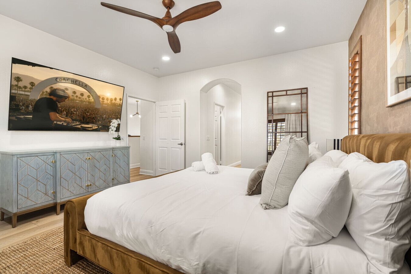 A bedroom with a large bed, a ceiling fan, a TV mounted on the wall, and a wooden headboard.