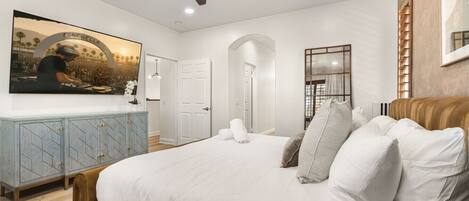 A bedroom with a large bed, a ceiling fan, a TV mounted on the wall, and a wooden headboard.