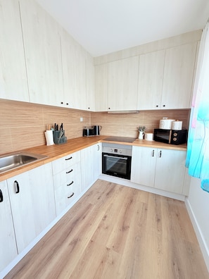 Una cocina moderna con armarios de madera clara, un lavamanos de acero inoxidable, horno empotrado y microondas.
