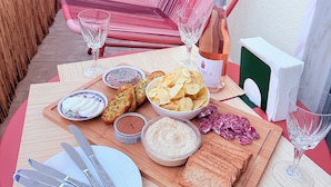 Une table dressée avec de la nourriture et des boissons, notamment du pain, des chips et une bouteille de vin, sur un balcon agrémenté d’une chaise en osier et d’une clôture en bois en arrière-plan.