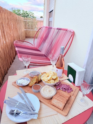 Una mesa dispuesta con comida y bebidas, incluyendo pan, patatas fritas y una botella de vino, en un balcón con una silla de mimbre y una valla de madera al fondo.