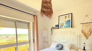 Une chambre à coucher avec un lit, un luminaire suspendu, une table de chevet, un tableau accroché au mur et une vue par la fenêtre.