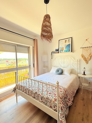 Un dormitorio con cama, lámpara colgante, mesita de noche, cuadro en la pared y vista por la ventana.
