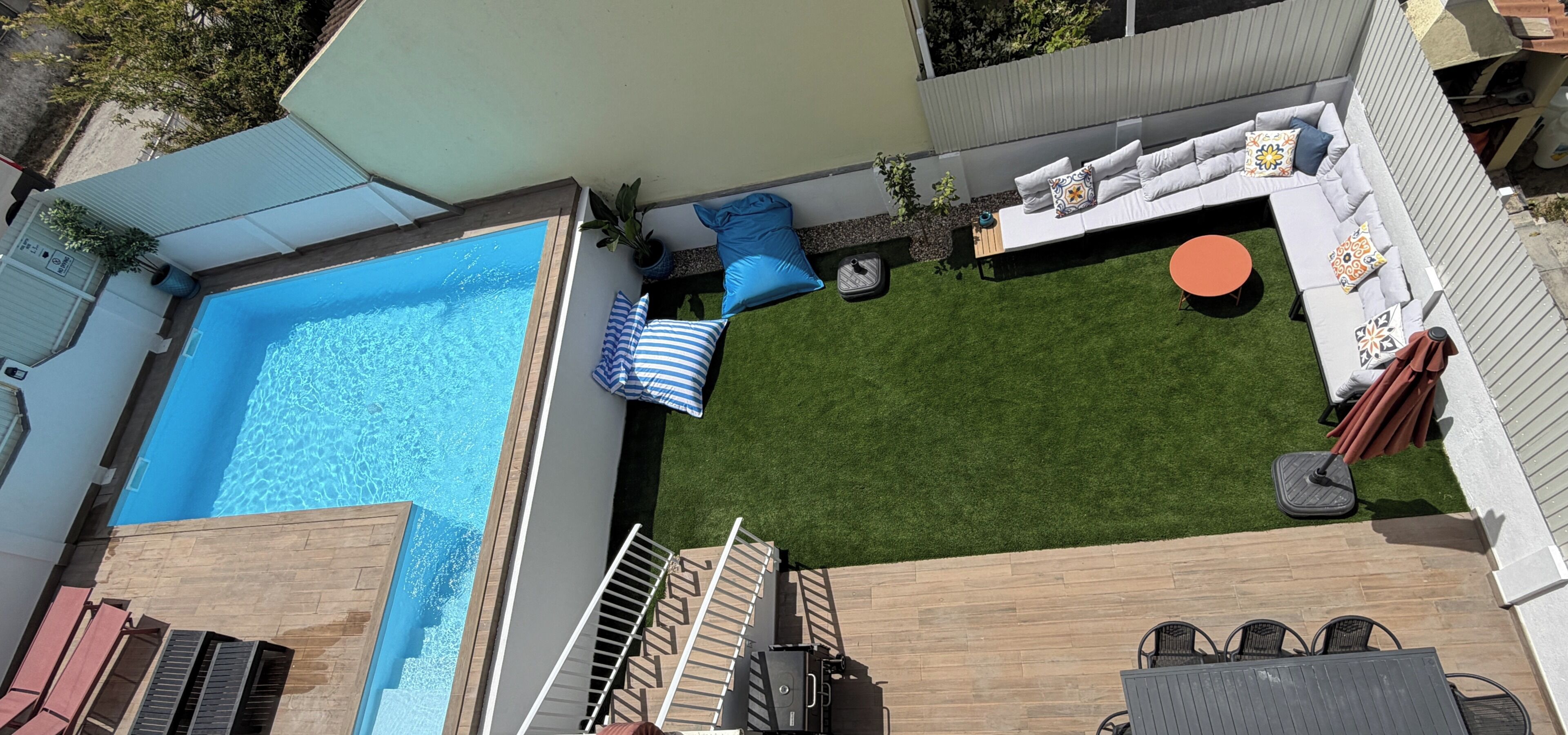 A backyard with a swimming pool, a grassy lawn, a seating area with a sofa and chairs, and a wooden deck.