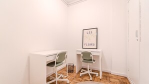 Ein kleiner, sauberer Raum mit zwei weißen Schreibtischen, grünen Stühlen, einem gerahmten Bild an der Wand und einem Teppich auf dem Boden.
