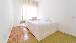 Ein helles Schlafzimmer mit Holzboden, einem Bett mit weißer Bettwäsche, einem Nachttisch und einem Fenster mit Jalousien.