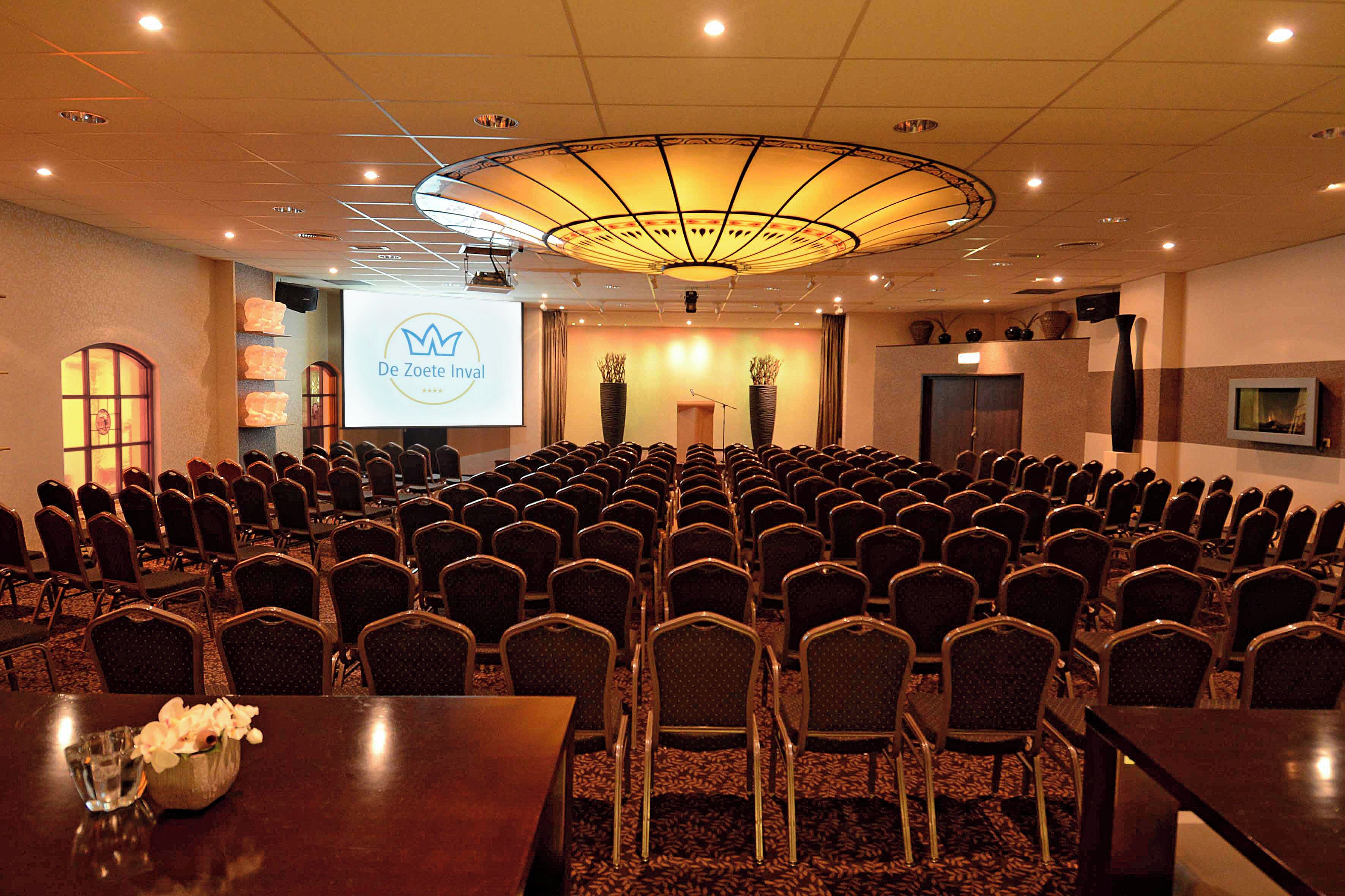 Sala de conferências com fileiras de cadeiras, um púlpito e uma tela exibindo 'De Zoete Inval'.