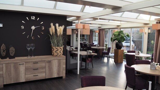A modern dining area with a black wall clock, wooden furniture, and large windows. - De Zoete Inval (Haarlemmerliede)