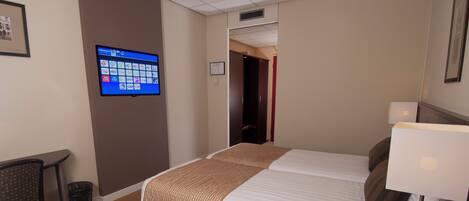 A hotel room with a bed, a desk with a lamp, a chair, a TV on the wall, and a trash can.