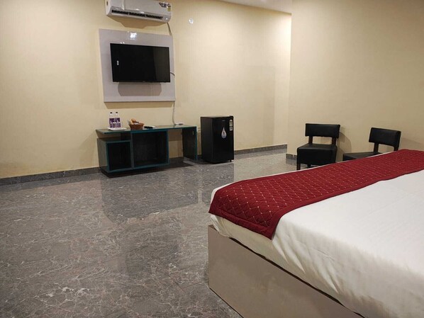 A hotel room with a bed, a TV mounted on the wall, a small refrigerator, a desk with a computer and some items, and two chairs. - The Aamantran Greens (Noida)
