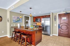 A modern kitchen with a central island, stainless steel appliances, wooden bar stools, and pendant lighting.