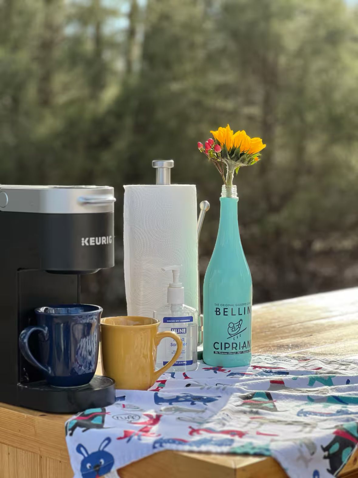 A coffee maker, two mugs, a tissue roll, a bottle of hand sanitizer, and a vase with flowers on a table with a view of trees.