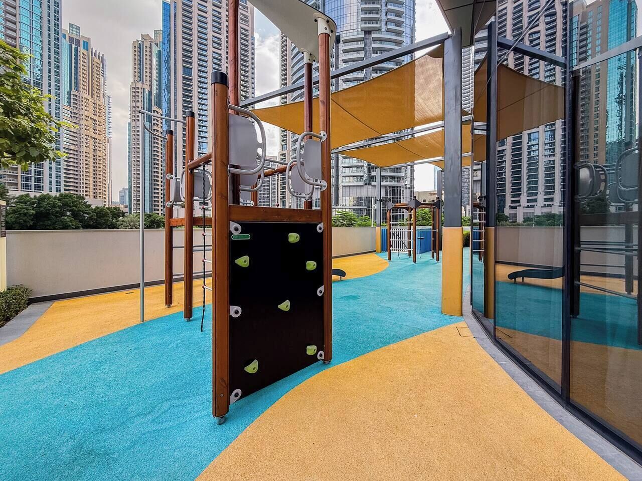 Een buitenspeelplaats met klimconstructies en een blauwe en gele rubberen bodem, omringd door hoge gebouwen.