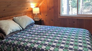 Quarto de cabana de madeira com cama, almofadas, mesa de cabeceira com abajur e janela com vista para árvores.