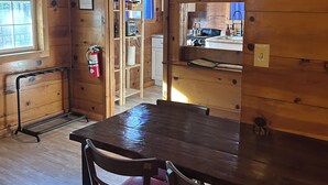 Interior de uma cabana de madeira, com uma mesa de jantar e cadeiras, área de cozinha visível através de uma porta aberta e um extintor de incêndio fixado na parede.