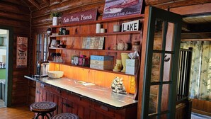A rustic cabin interior with a wooden bar area, shelves with various items, and a green kitchen in the background.