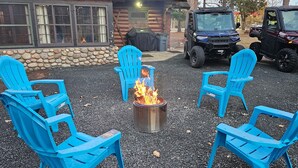 A fire pit with blue chairs arranged around it, a stone wall, and a building with windows in the background.