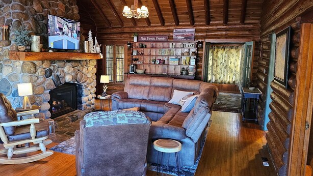 A cozy log cabin interior with a stone fireplace, leather sofas, a television, and a bookshelf.