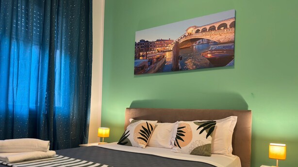 A hotel room with a bed, two bedside tables, a nightstand, a lamp, a window with blue curtains, and a wall painting of a bridge. - United Residence (Mestre)